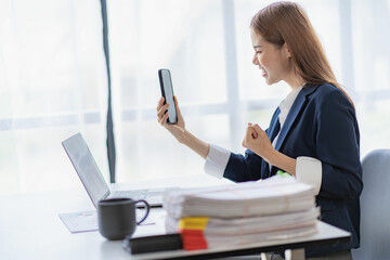 Successful young Asian businesswoman uses smartphone and laptop to work in office with financial graph document on desk.