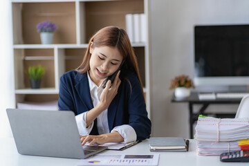Successful young Asian businesswoman uses smartphone and laptop to work in office with financial graph document on desk.