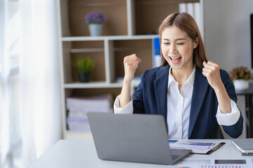 Happy young Asian businesswoman in a successful business excitedly raising her hand with a laptop...