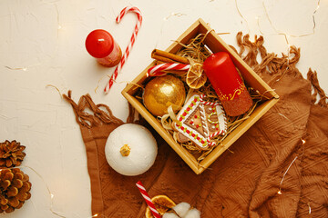 Flat lay of christmas gift box, blanket and decorations on white textured table. New Year's atmosphere. Candy cane, candle with ornate, gingerbread house, dried orange and cones. Festive still life.