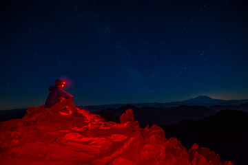 Athletic adventurous male hiker sitting on top of a mountain with a red headlamp shinning, looking at the stars and Mount Rainier.  © Pelo Blanco Photo