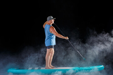 A man on a sub board with an oar in his hands on a black background in the fog.