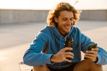 White young man drinking water while sitting on workout mat outdoors