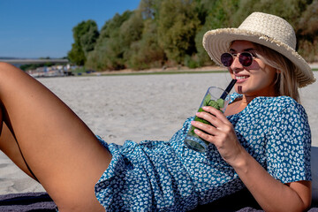 Beautiful young woman with a straw hat, lying on a deserted sandy beach, enjoying the sun and drinking cocktail.