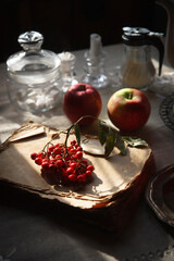 Obraz na płótnie Canvas A bunch of rowan lying on the old and scrubby book, two red apples and the other tableware for tea time on gray rustic table, autumn morning concept.