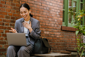 Asian woman in earphones working with laptop while sitting on bench