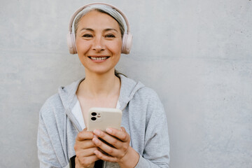 Fototapeta premium Asian mature woman using cellphone while standing by wall