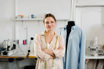 Portrait of a beautiful seamstress in glasses with a measuring tape and working in a textile factory near a manikin in a blue shirt. High quality photo