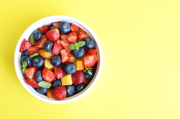 Fruit salad in a white cup on a yellow background. Sliced strawberries, peaches and blueberries with mint leaves. Free space for text.