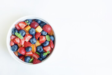 Fruit salad in a white cup on a white background. Sliced strawberries, peaches and blueberries with mint leaves. Free space for text.