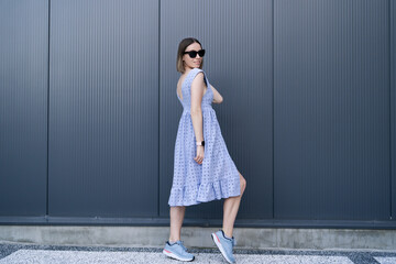 Young Caucasian Woman posing in blue dress on gray wall background outdoors. Female wearing sunglasses