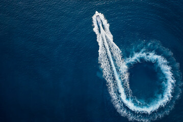 Aerial top view of a motor powerboat forming a circle of waves and bubbles with its engines over the blue sea - obrazy, fototapety, plakaty