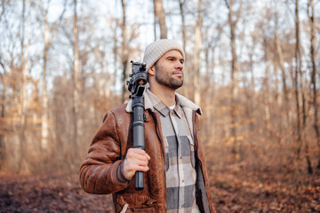 A portrait of a young white man posing in the woods dressed in autumn clothes: brown jacket, checkered canadian style shirt, off-white beanie, holding gimbal for videography