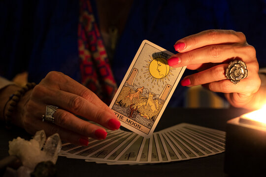 Fortune teller hands showing The Moon tarot card, symbol of intuition, during a reading. Close-up with candle light, moody atmosphere.