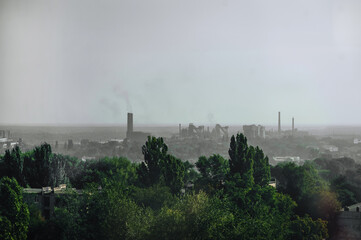 Green plants, trees grow against the backdrop of a factory with pipes and dirty smoke against the sky. Photography, industry.