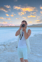 Obraz premium Adult man photographer taking photographs with digital camera at Dead Sea beach in Ein Bokek, Israel. Handsome muscular man using picture with modern dslr camera at the beach of seashore. Travel