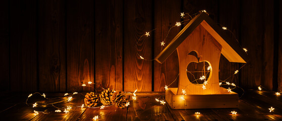 Christmas and New Year background. Wooden toy house feeder and pine cones in golden warm light of garland. Old wooden background.