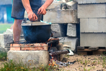 Defocus man cooking fish soup in the iron bowler over a campfire. Fish soup boils in cauldron at the stake. Soup in a pot in the fire. Camp at day. Nature green grass background. Out of focus - obrazy, fototapety, plakaty