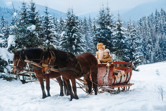 Two children ride on a retro sleigh with horses in winter time. Mountain snowy landscape background. Tours, horse riding.