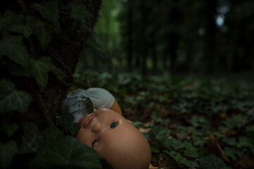 Close up dark scene of scary child doll in deep woods growth by ivy everywhere just before night. Restless low light scene creating spooky feeling suitable for Halloween holiday or some cemetery story