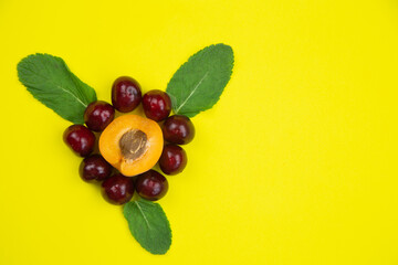 A creative flower consisting of half an apricot with a heart stone and petals in the form of a cherry, decorated with mint leaves lies on the side on a bright yellow background with a place for text