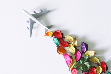 A light-colored toy airliner with a contrail of multi-colored flower petals. Concept of eco-friendly biofuel and green revolution. White background. Copy space.