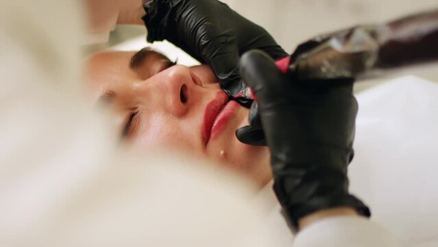 Permanent makeup procedure applying rose red pigment using tattoo machine in beautician salon. Microblading tattoo lips