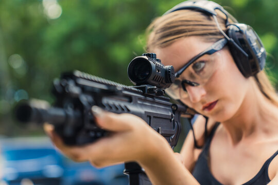 Closeup shot of a loupe of a black rifle held by focused powerful caucasian woman in protective shooting range gear. Blurred background. High quality photo