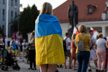 In focus: migrant blond woman stands in crowd (square) and covers shoulders by Ukrainian blue and yellow flag. Back view. Horizontal plane