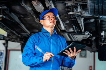 Auto mature mechanic repairman in uniform using digital tablet checking auto suspension repair in the garage, change spare part, check the mileage of the car, checking and maintenance service concept.