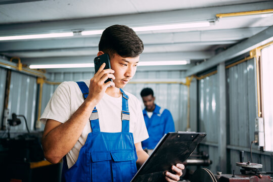 Auto mechanic repairman in uniform talking on smartphone with the customer in the garage and looking on the clipboard while colleague working in the background, repair car brake system service center.