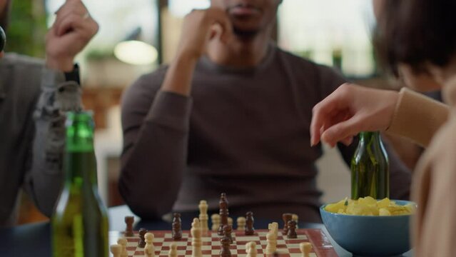 Cheerful man playing board games with group of friends, enjoying chess match competition at home. Young adult having fun with people, drinking beer and eating snacks. Handheld shot. Close up.