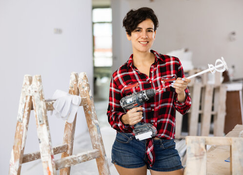 Positive young brunette woman in denim shorts and plaid red shirt doing renovations in her apartment, standing near wooden stepladder with paint mixer in hands ..