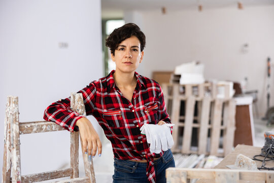 Positive relaxed young brunette standing leaning on paint-smeared wooden stepladder, holding work gloves in apartment under renovation..