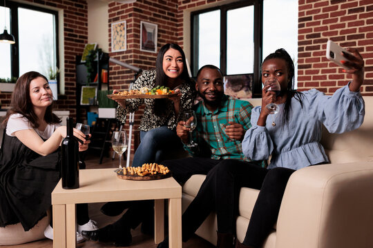 Multiracial cheerful young people at home celebrating birthday while taking selfie photos together and drinking wine. Group of friends drinking alcoholic beverages while photographing themselves.