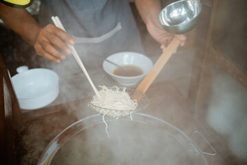 hands of seller drain boiled noodles with a sieve and chopsticks from the pan on the cart