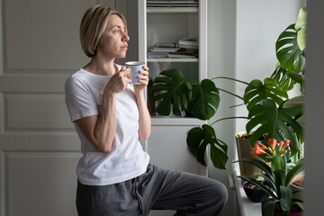 Tranquil middle-aged female enjoys hot drink from mug standing at windowsill. Mature blonde woman...