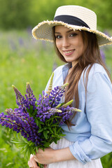 Beautiful young girl in a white dress, straw hat with a bouquet of violet flowers in her hands and picnic basket. Pretty woman in summer in the blooming field holding a bunch of purple lupin