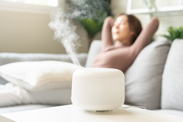 Modern air humidifier during relax or rest, happy blurred asian young woman, girl enjoying aromatherapy steam scent from essential oil diffuser comfortable in living conditions room,apartment at home - obrazy, fototapety, plakaty