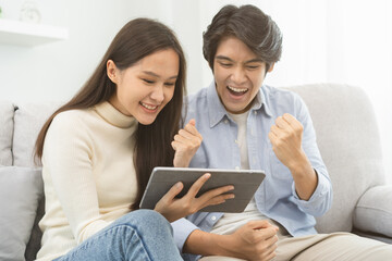 Fototapeta na wymiar Happy excited, smiling asian young couple love using tablet pc, great deal or business success, received or getting cash back, tax refund, good news by mail while sitting on sofa, couch at home.