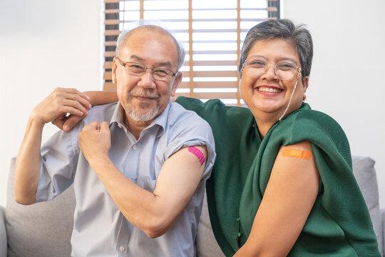Coronavirus Vaccination, happy asian elderly, aged family smile strong together, showing bandage on arm with protect of covid-19 after injection of vaccine, sitting on couch in living room at home.
