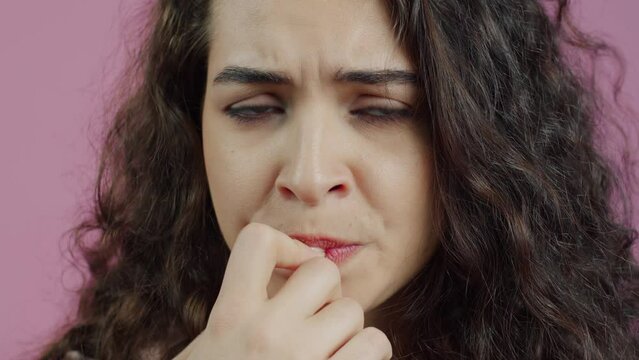 Close-up portrait of nervous young woman biting finger nails and looking at camera against pink color background. People and emotions concept.
