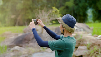 Asian man Wear a hat and bag strap , standing on the ground, in the middle of a rice field,He raised his mobile phone with his hand, took a picture of a green rice field, kept it in his phone.