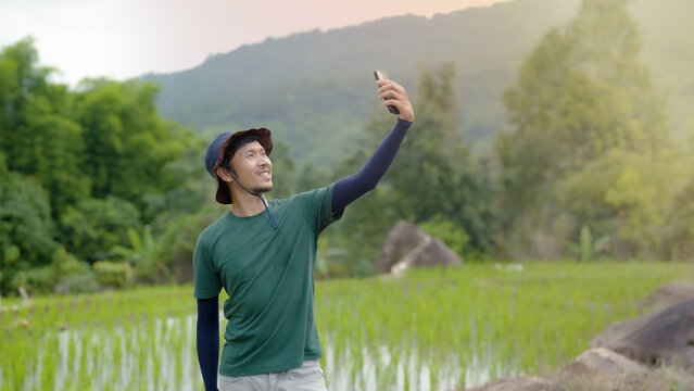 Good looking Asian guy, grows a mustache, wears his favorite sun hat,He picked up his cell phone to take pictures, the beautiful green fields, in front of him,keep,happy face.
