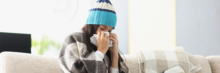 Sick woman in warm hat blowing her nose into paper napkin at home