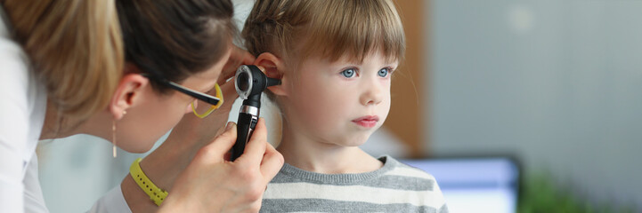 Female pediatrician looking at ear of little girl using otoscope in clinic