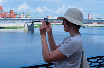 Obraz premium Blue-color toned photography of woman tourist in a straw hat makes a mobile photo on a city street