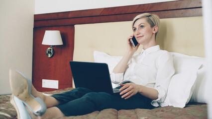 Smiling blonde businesswoman in white shirt using on laptop and talking at mobile phone while lying...