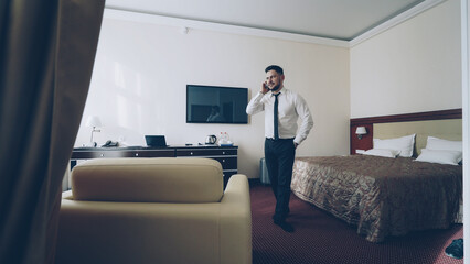 Confident young businessman talking on mobile phone while walking around hotel room. Travel,...