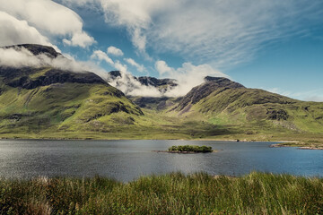 Mountains of Connemara, beautiful nature scene in Ireland with small lake with island and green tall mountains in low clouds. Warm sunny day. Travel and sightseeing. Irish landscape and nature.